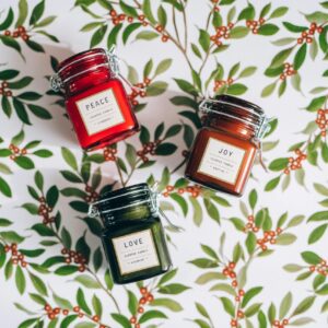 Collection of scented candles labeled Peace, Joy, Love on a leafy holiday background.